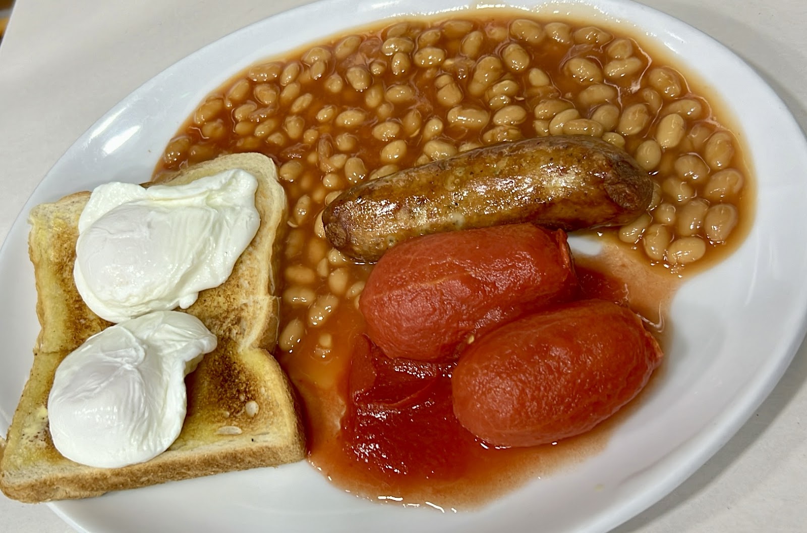 A breakfast plate from above: two poached eggs on white toast, a browned sausage, two grilled tomato halves, and a generous pool of baked beans, on a plain white ceramic plate