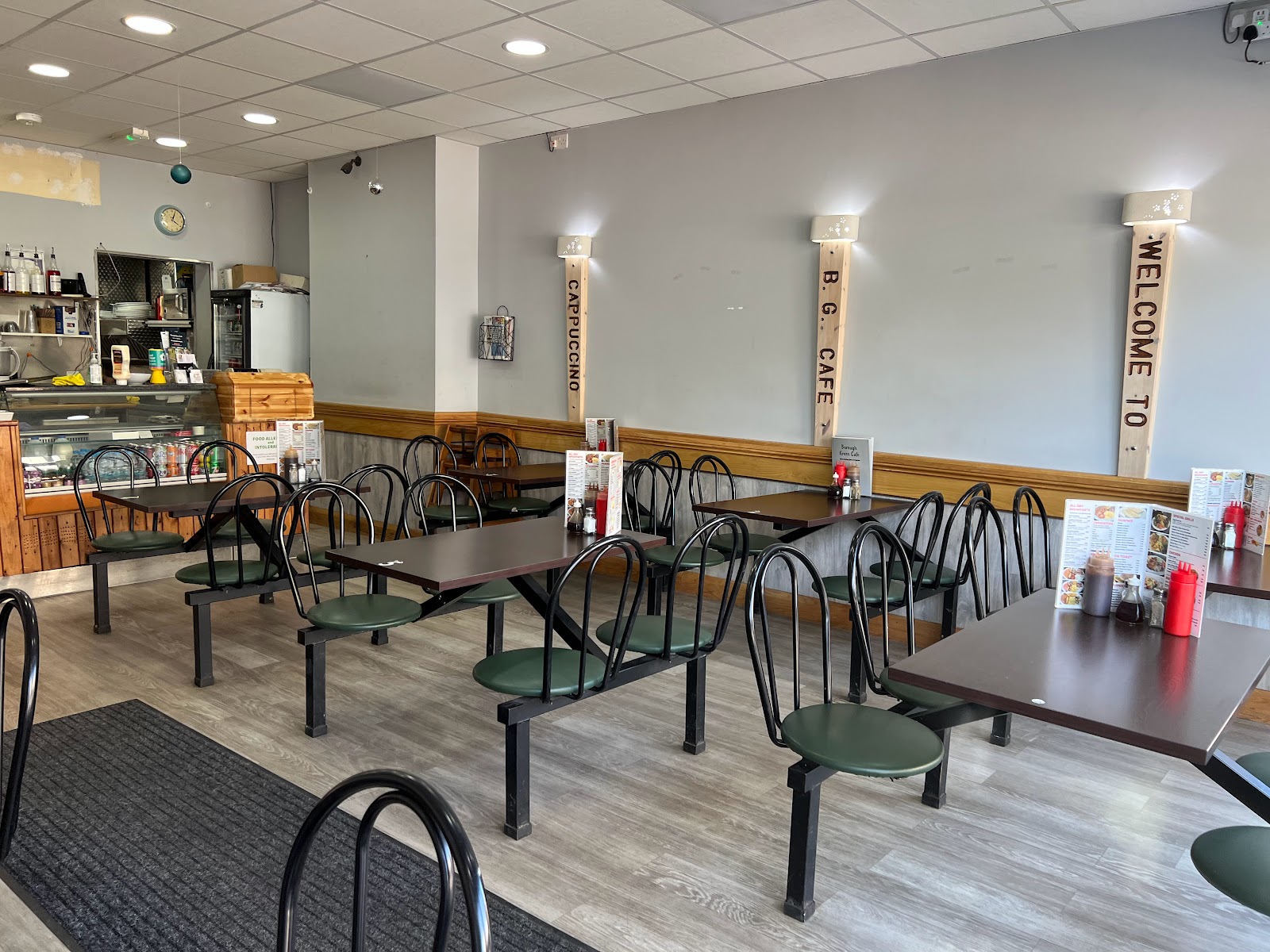 Inside Borough Green Cafe, three wooden columns hand-painted with the words Welcome To, B.G. Cafe, and Cappuccino in white block letters running vertically, bright open room with white walls and grey paint dado, brown laminate tables, black metal chairs with green vinyl seats arranged in fixed booths, stainless service counter at the back