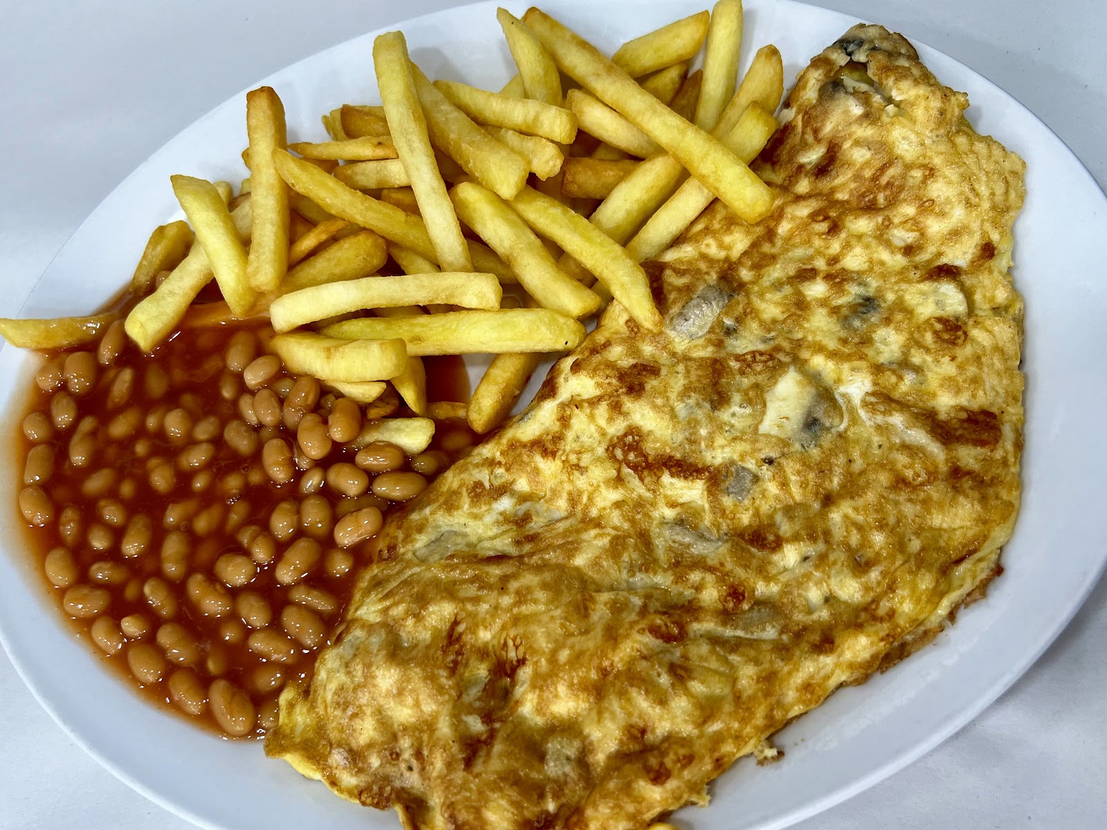 A folded mushroom omelette on a white plate with a generous pile of golden chips and a pool of baked beans on the side, lunchtime portion