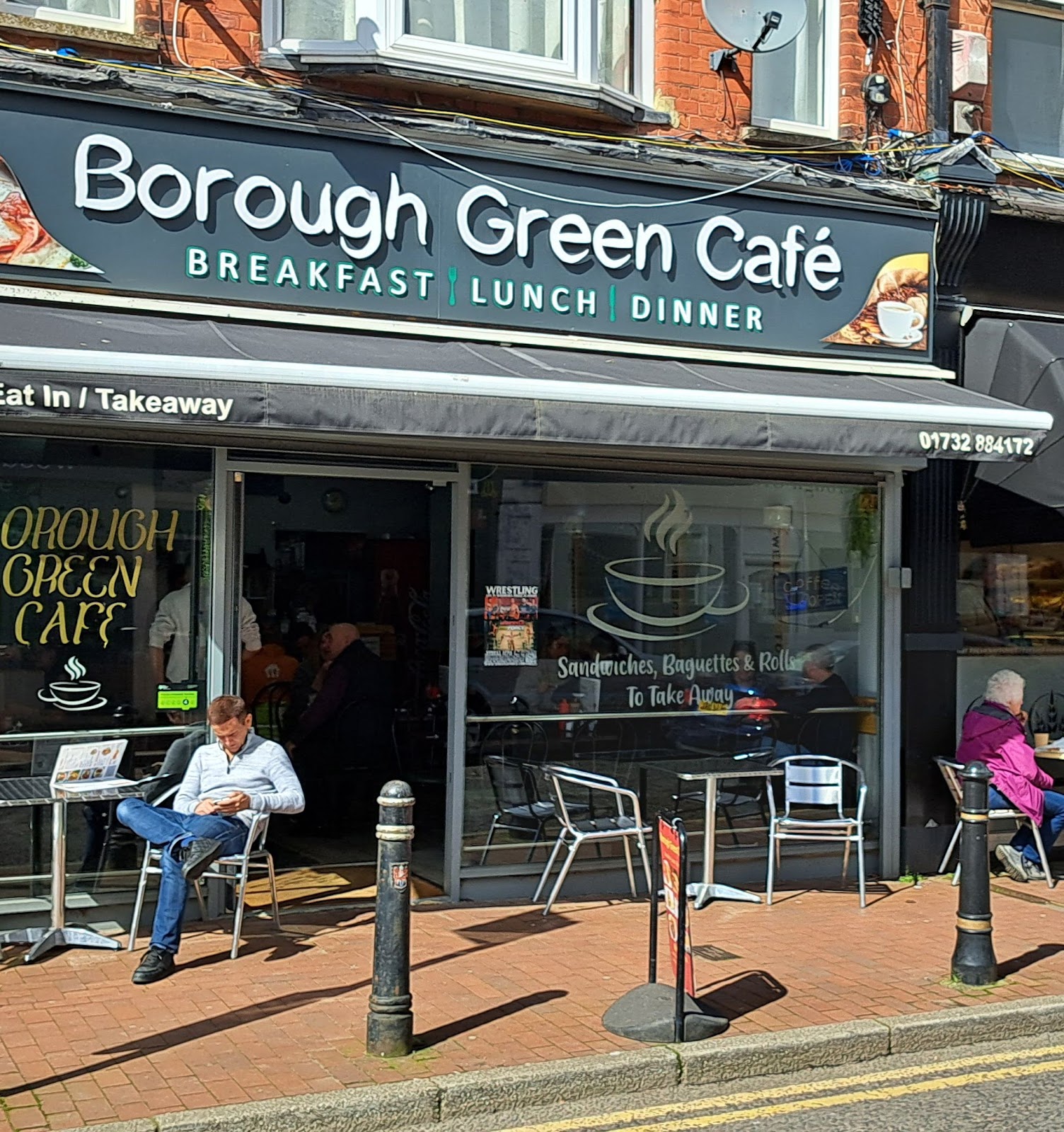 Borough Green Cafe from across the high street, black fascia with the cafe name in white serif and Breakfast Lunch Dinner underneath, chrome cafe chairs on the pavement with a customer reading at a small table, brick parade with shops either side